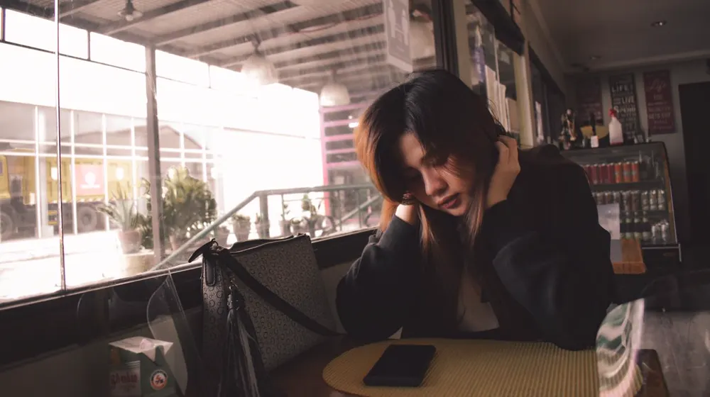 Illustrative photo of a woman sitting alone at a cafe table with her phone nearby